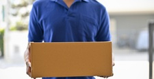 A cropped shot of a delivery man in a blue shirt holding a cardboard box in front of the door.